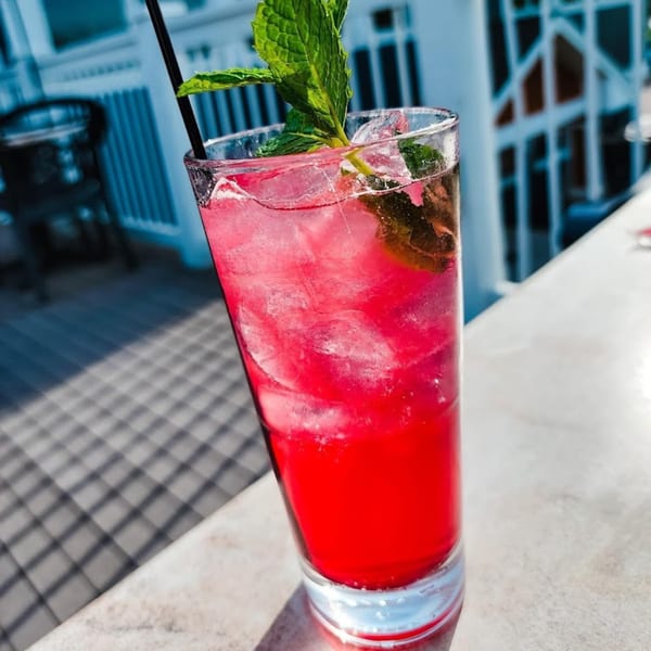 A vibrant red cocktail with ice, a black straw, and fresh mint sits on a sunlit table.